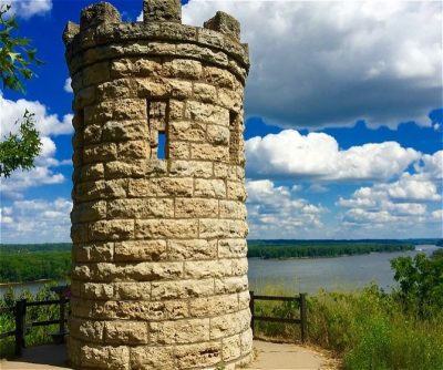 Stone lookout tower with window, overlooking a river surrounded by greenery under a blue sky with fluffy clouds.