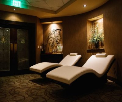 Relaxing spa lounge with two white chaise lounges, soft lighting, and decorative plants in a warm-toned setting.