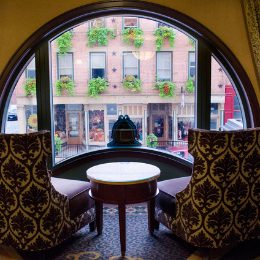 Cozy hotel seating area with two plush chairs and a circular window overlooking a vibrant street with flowered balconies.