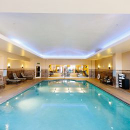 Indoor hotel pool with a turquoise blue water, loungers, under soft lighting.