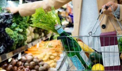 A person holds a shopping basket filled with fresh vegetables, including leafy greens, cucumbers, and a lemon, amidst various produce in a grocery store aisle.