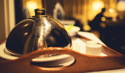 Room service tray with a silver cloche covering a meal and two cups, set in a softly lit hotel room.