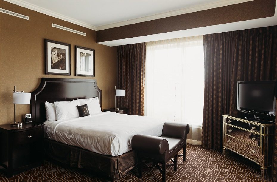 Cozy hotel room featuring a queen bed, dark wood furniture, and large windows with sheer curtains. Includes a flat-screen TV.