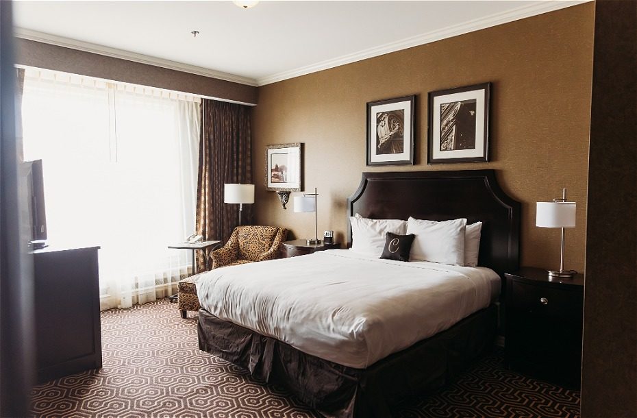 Cozy hotel room with a king-size bed, armchair, and large windows draped in sheer curtains. Warm, earthy tones throughout.