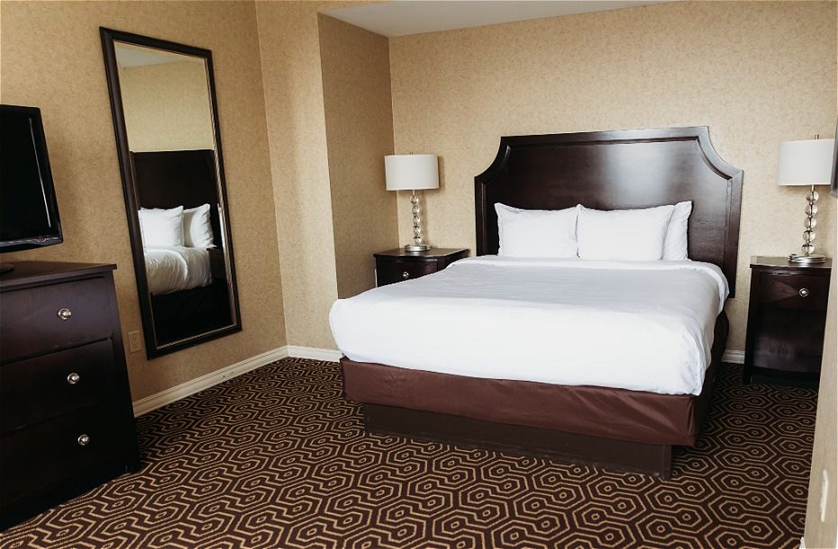 Contemporary hotel room featuring a queen bed, bedside tables, a TV, and a large mirror on a patterned carpet.