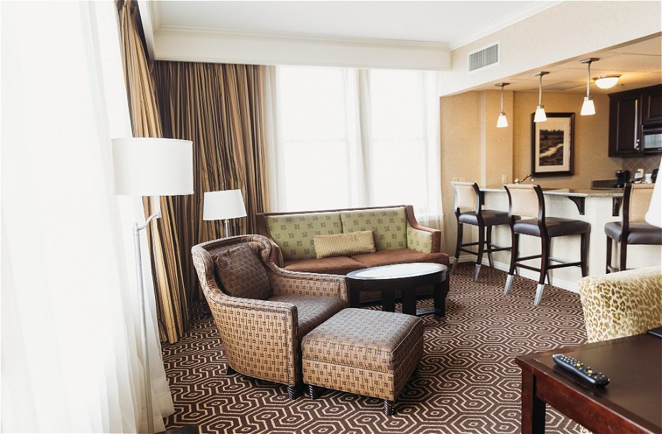 Spacious hotel living area with a sofa, armchairs, coffee table, and bar seating. Bright natural light from large windows.