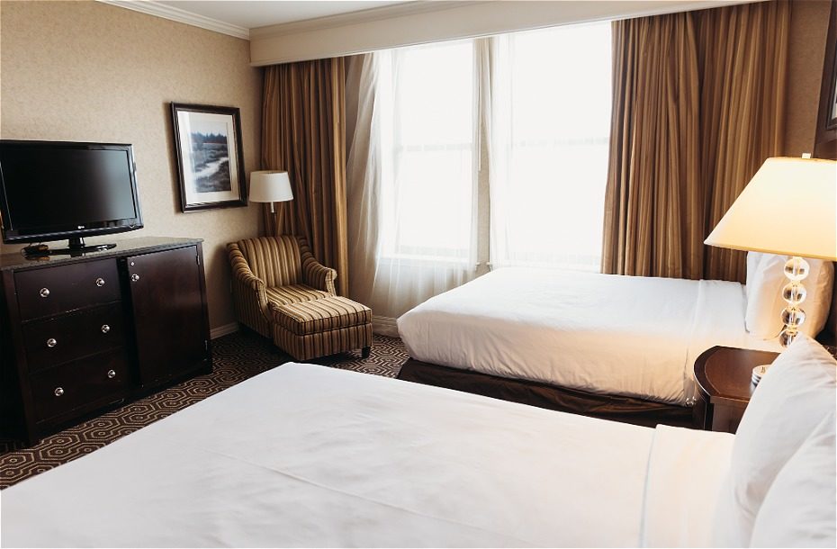 Modern hotel room featuring two double beds, a television, and a cozy armchair by large windows with natural light.