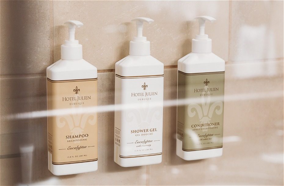 Three luxury toiletries: eucalyptus shampoo, shower gel, and conditioner in pump bottles on a tiled bathroom wall.