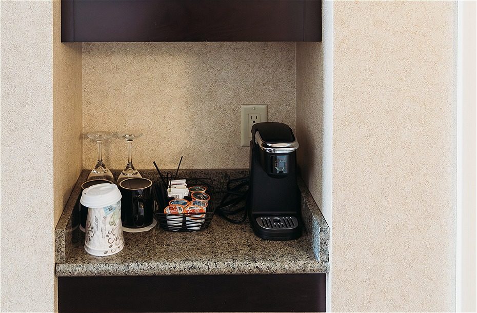 Coffee station with a coffee maker, cups, creamer, and glasses on a granite countertop in a hotel room.