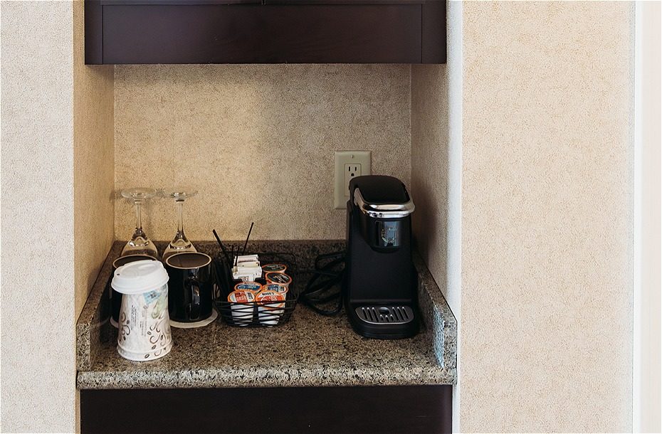 Coffee station with a coffee maker, cups, creamers, and glassware set on a granite countertop in a hotel room.