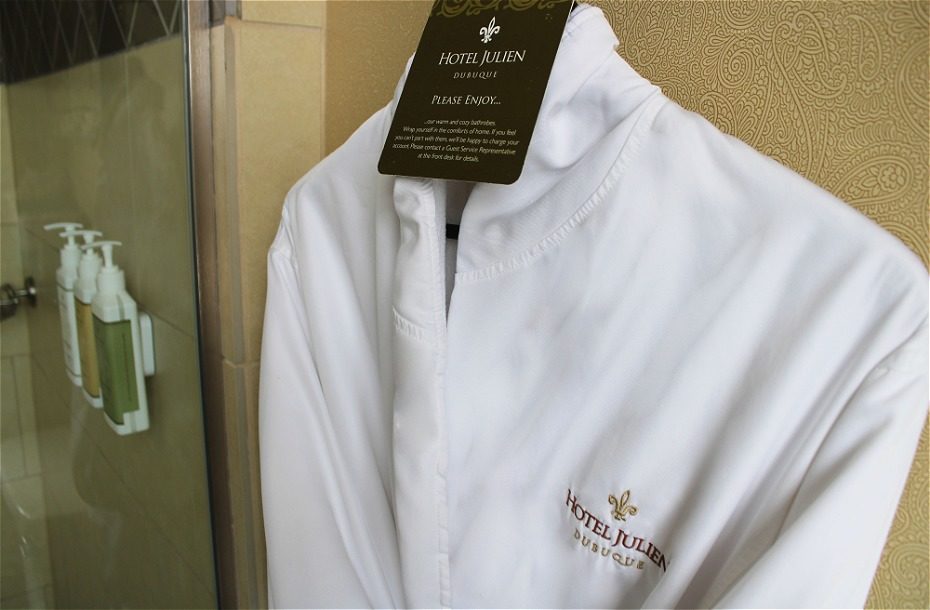White hotel bathrobe hanging on a hook, with lotion bottles visible in a bathroom setting.