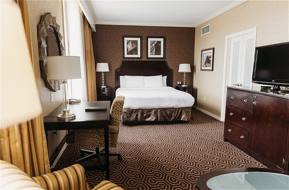 Cozy hotel room featuring a king bed, desk, TV, and patterned carpet, with natural light from large windows.