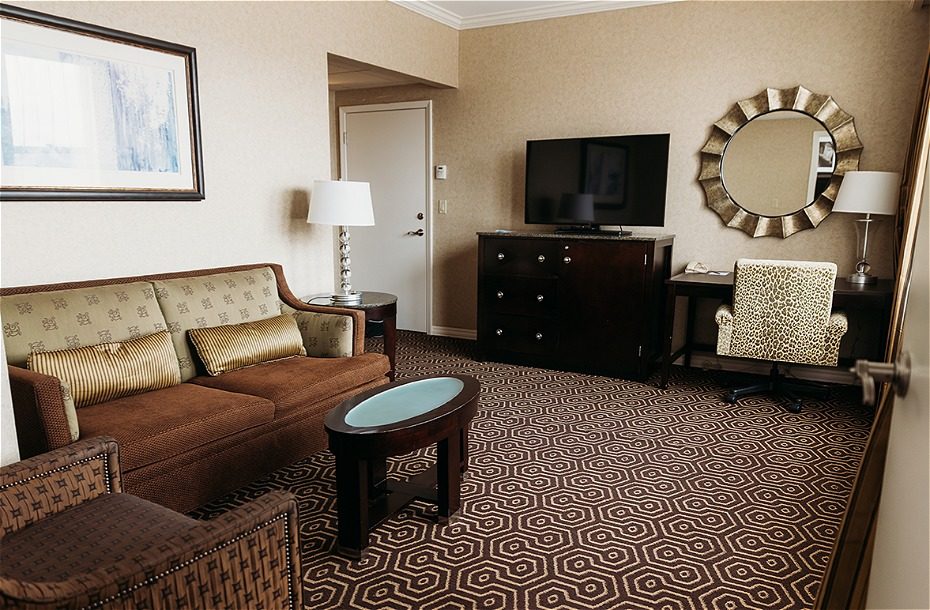 Spacious hotel room featuring a seating area with a sofa, modern TV, desk, and patterned carpet. Bright and inviting decor.