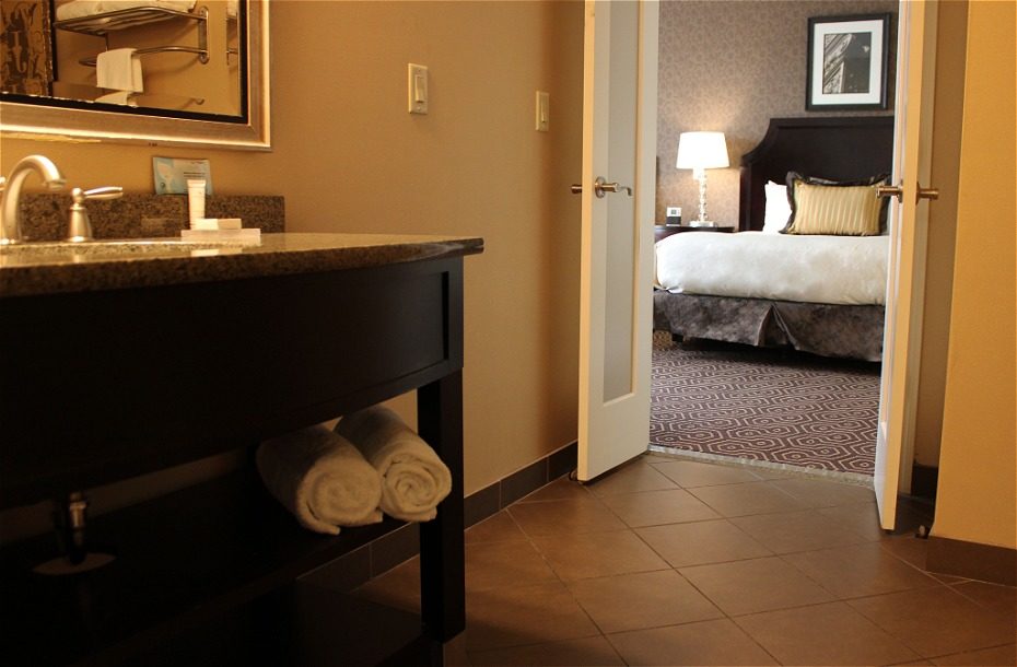 Bathroom view with towels stacked, leading to a well-furnished hotel room featuring a bed and bedside lamp.