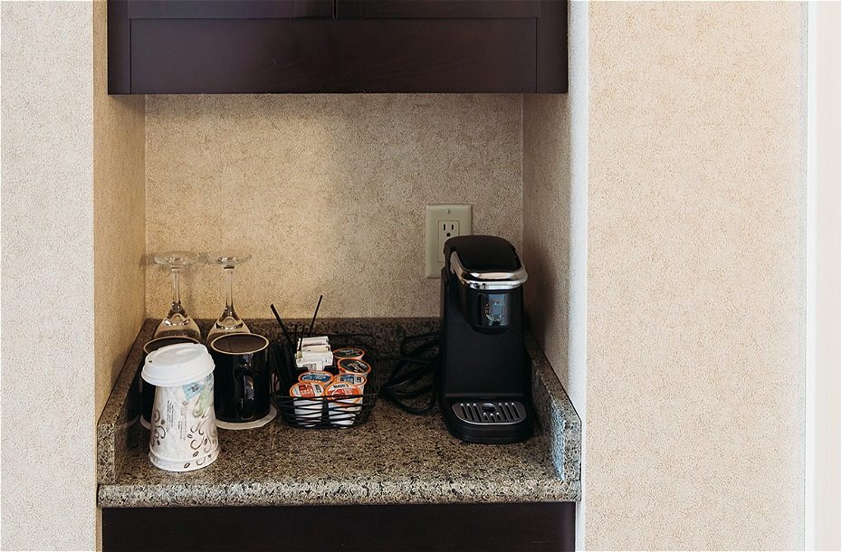 Coffee station with a coffee maker, mugs, creamers, and glasses on a granite countertop in a hotel room.