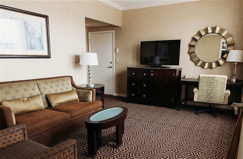 Spacious hotel suite with a comfortable sofa, TV, stylish chair, and decorative mirror on patterned carpet.