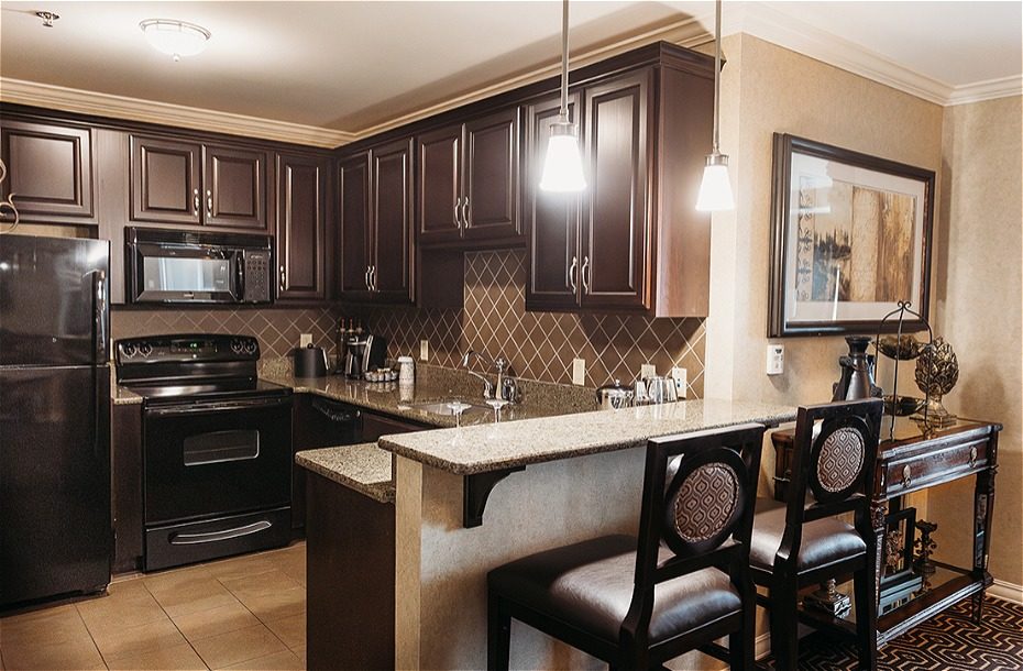 Modern kitchenette with dark wood cabinets, granite countertops, and seating for two in a hotel room setting.