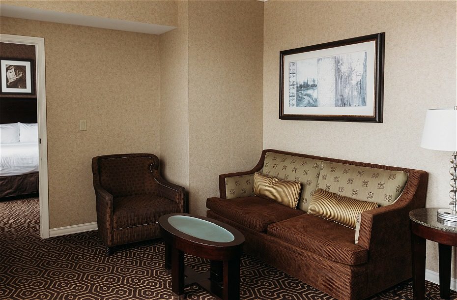 Cozy hotel seating area with a sofa, armchair, round table, and tasteful artwork on the wall. A bedroom is visible in the bac