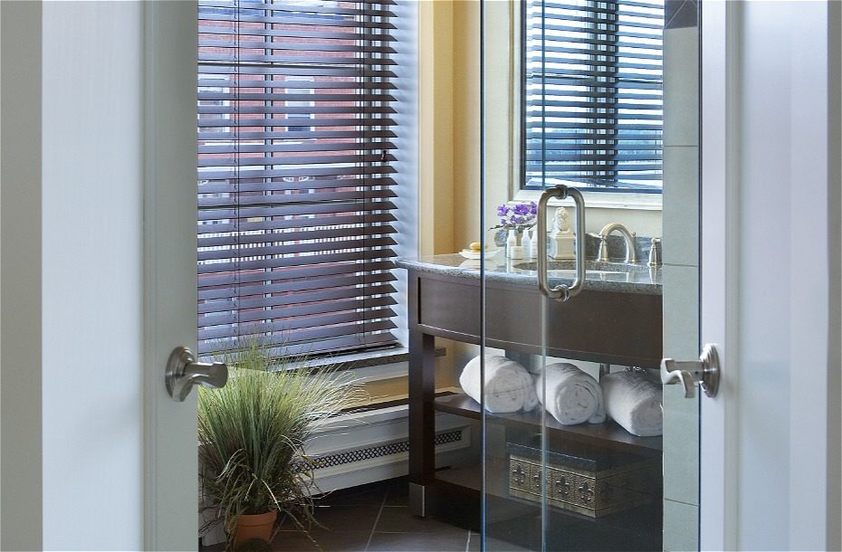 Luxurious hotel bathroom featuring a glass shower, modern vanity, and neatly stacked white towels by a window with blinds.