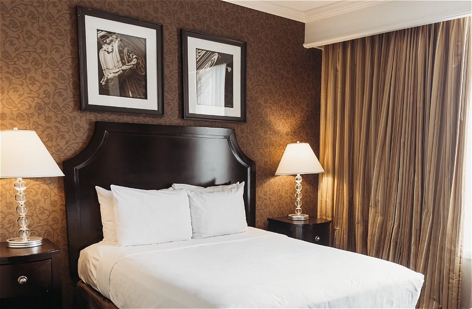Cozy hotel room with a queen bed, framed artwork, and elegant curtains. Includes bedside lamps and a dark wood nightstand.