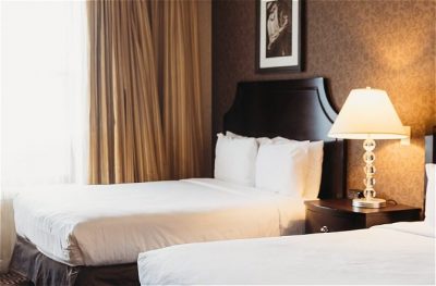 Cozy hotel room featuring two queen beds, warm lighting, and rich brown wall decor. A lamp and artwork add elegance.