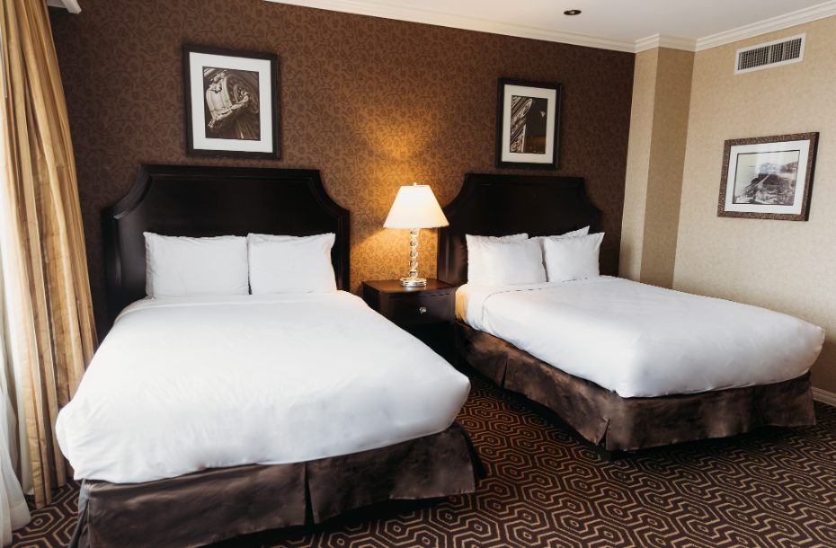 A cozy hotel room featuring two queen beds, a bedside lamp, and decorative prints on textured brown walls.