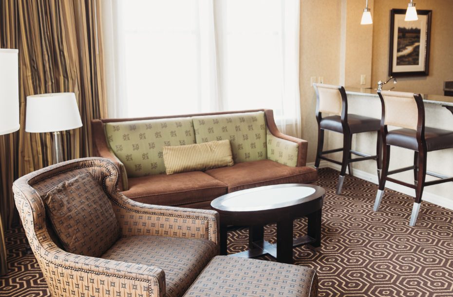 Spacious hotel living area featuring a sofa, armchair, coffee table, and bar seating, with ample natural light.