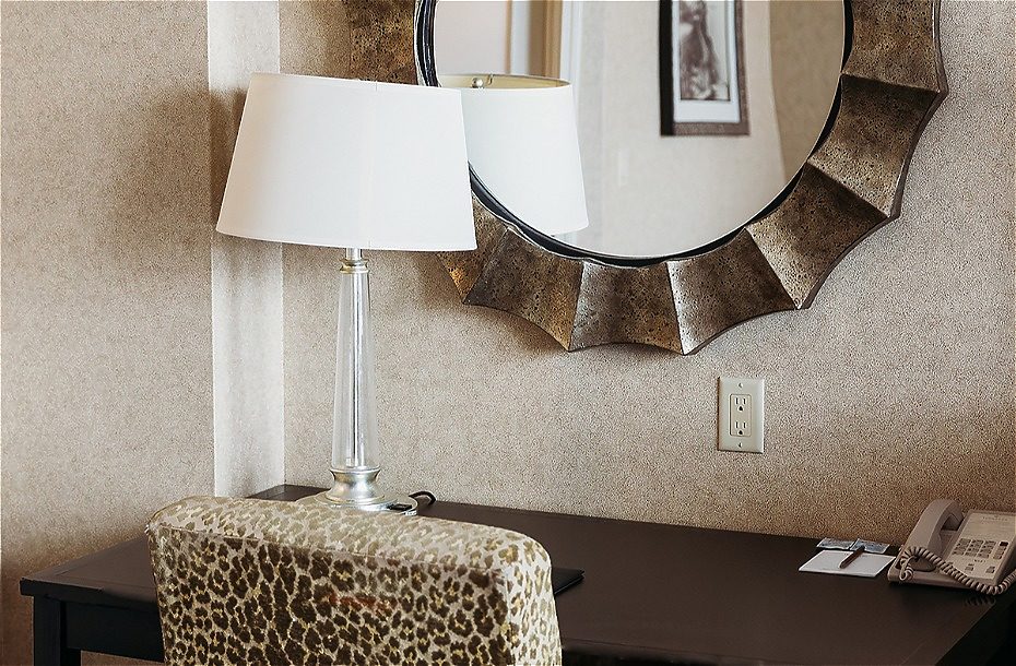 Stylish hotel desk area featuring a patterned chair, lamp, round mirror, and phone against a textured wall.