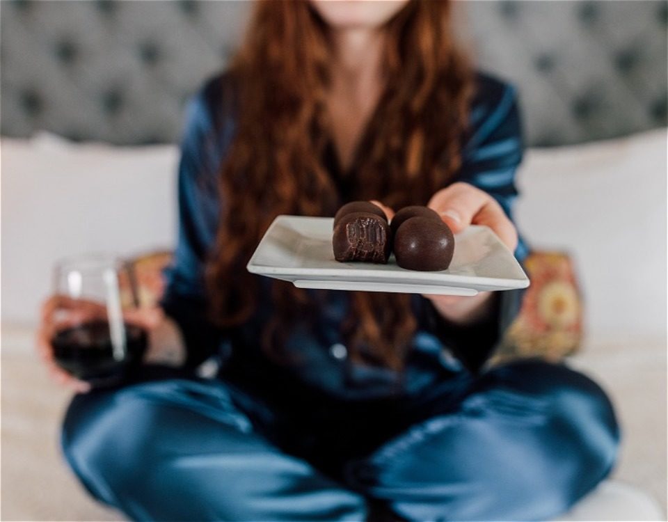Woman in silk blue pajamas sitting on a bed and holding a glass of wine while handing over a plate of chocolate truffles
