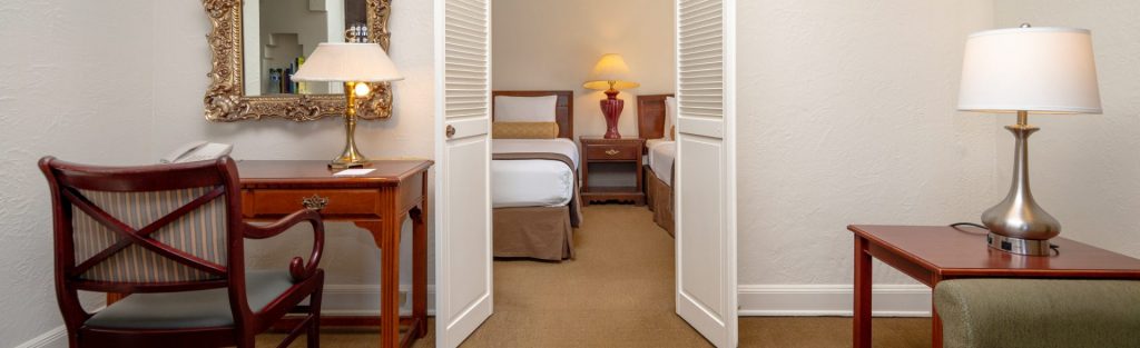 A wide view of a hotel room with a desk, chair, lamp, mirror, and bed.
